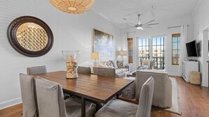 A modern living room with a wooden dining table, grey upholstered chairs, a large round mirror, and a ceiling fan. - The Newcastle Updated 3BR/2BA ~ Best Location! (Inlet Beach)