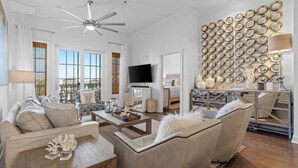 A spacious living room with a ceiling fan, a large TV, and a wall decorated with circular mirrors. - The Newcastle Updated 3BR/2BA ~ Best Location! (Inlet Beach)