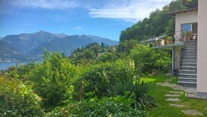 A lush garden with a stone pathway leading to a building with a balcony overlooking a lake and mountains.