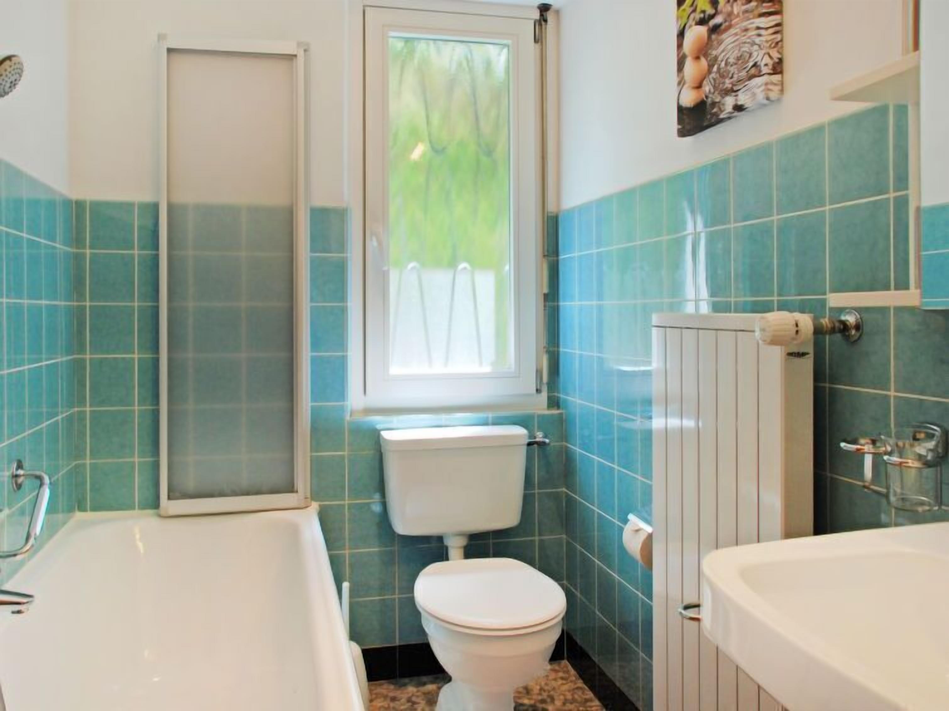 Ein Badezimmer mit Toilette, Badewanne und Waschbecken, ausgestattet mit blauen Fliesen und einem Fenster.