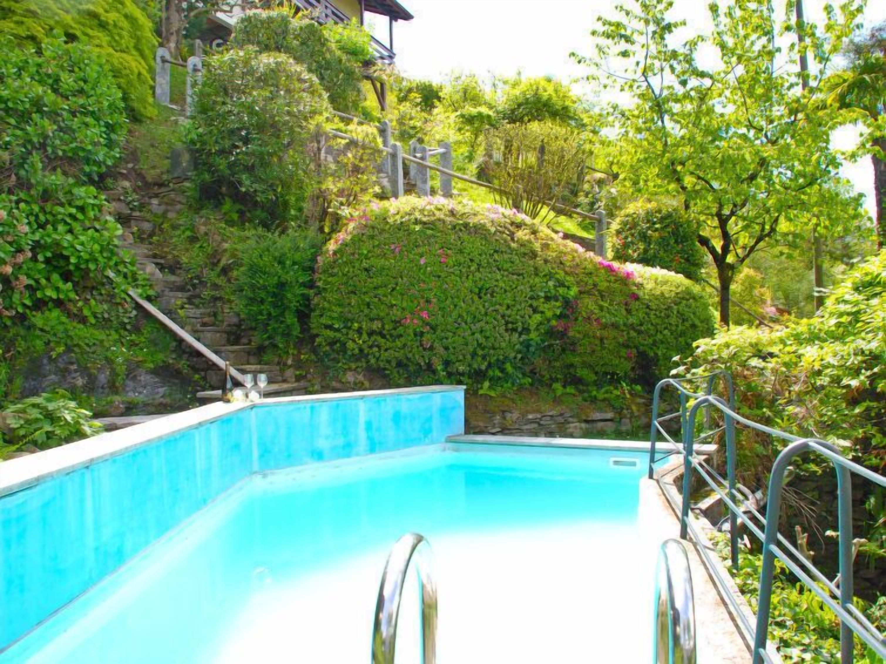 A swimming pool with a stone wall and metal railings, surrounded by lush greenery and a staircase leading up.