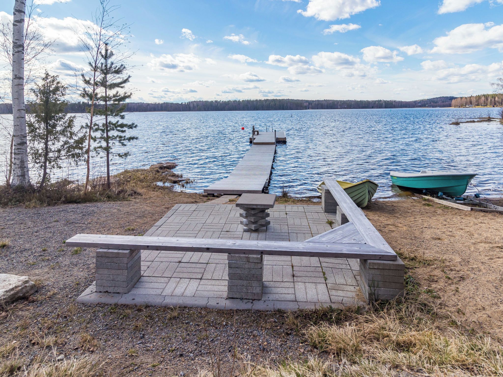 A lakeside wooden deck with a stone table and benches, a dock extending into the water, and a rowboat.
