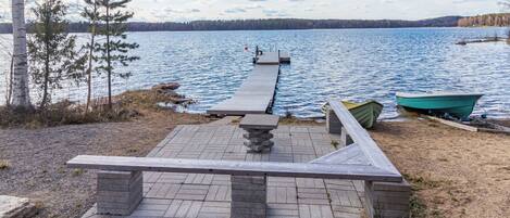 A lakeside wooden deck with a stone table and benches, a dock extending into the water, and a rowboat.
