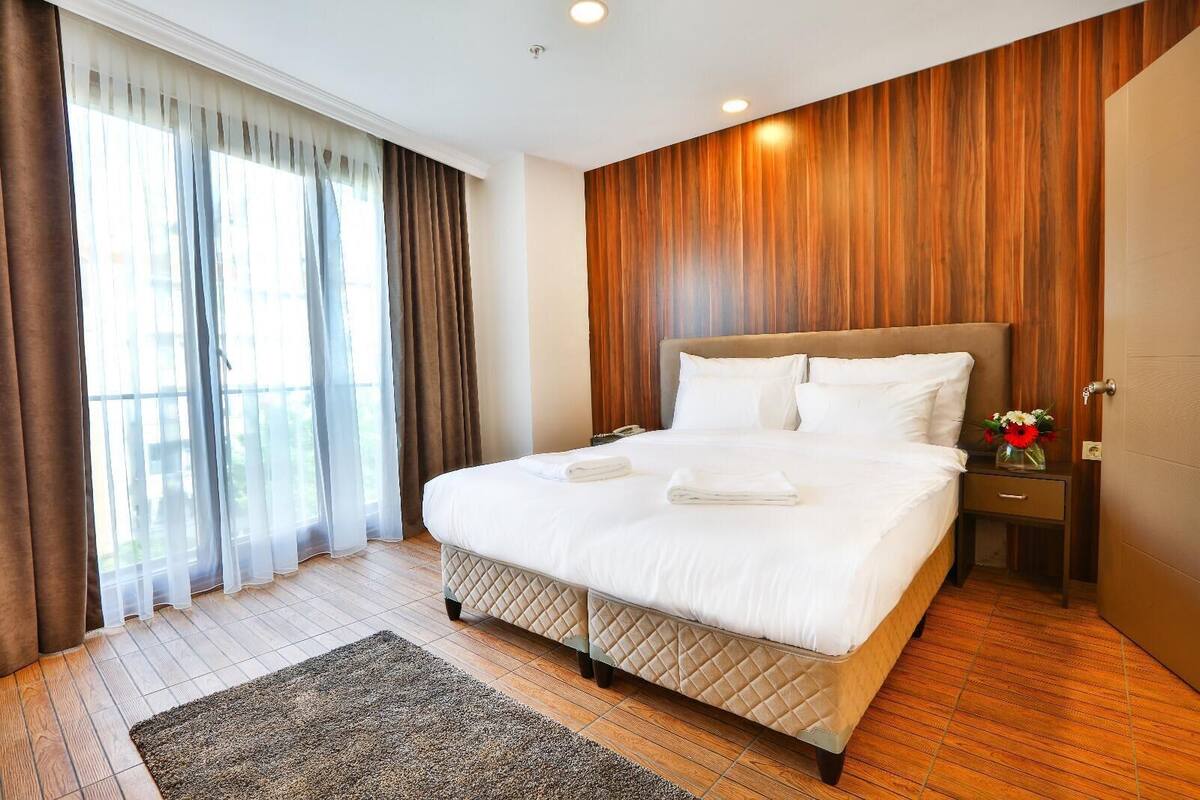 A hotel room with a large bed, wooden panel walls, a nightstand with a vase of flowers, and a view through sheer curtains.