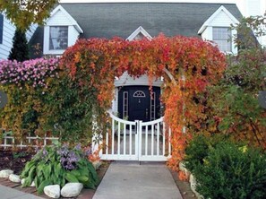 Une maison dotée d’un porche, d’un portail blanc et d’un jardin où poussent différentes espèces de plantes.