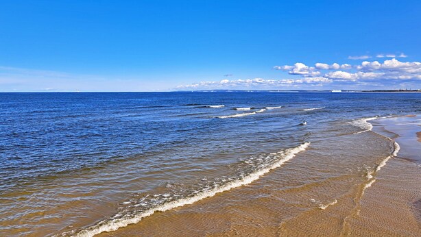 Ein Strand mit klarem, blauem Wasser, Wellen und einem Schiff in der Ferne.
