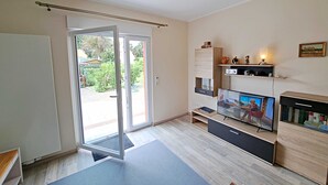 Ein modernes Wohnzimmer mit einer Schiebeglastür, die zu einem Garten führt, einem Flachbildfernseher, einem Holzschrank und einer kleinen Ablage mit Dekorationselementen.