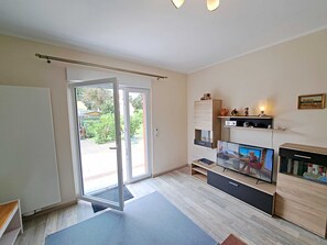Een moderne woonkamer met een schuifdeur van glas naar een tuin, een flatscreen televisie, een houten kast en een klein plankje met decoratieve objecten.