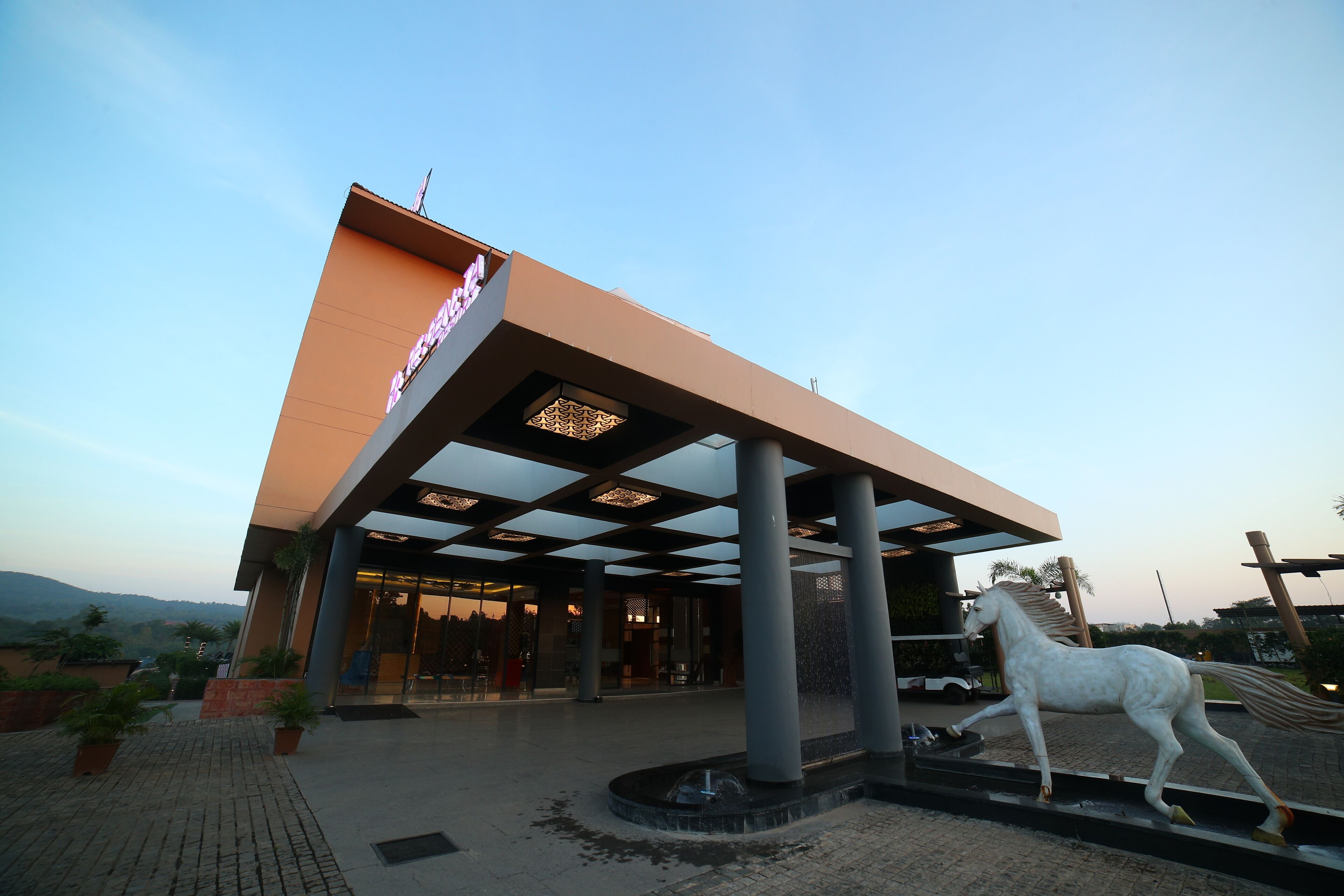 A modern building with a large entrance, a horse statue, and a landscaped area with potted plants.