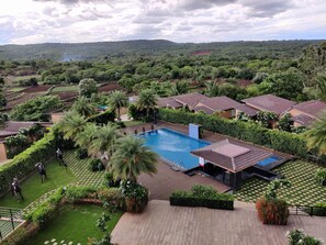 A resort with a swimming pool surrounded by lush greenery and palm trees, set against a backdrop of hills.