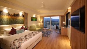Ein modernes Hotelzimmer mit einem großen Bett, einer Couch, einem kleinen Tisch und einem Fernseher. Das Zimmer hat Holzböden und ein großes Fenster mit Blick auf die Stadt.