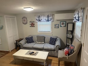 Een woonkamer met een geruite bank, een witte salontafel en een beige fauteuil. Er is een raam met zonwering, een wandklok en decoraties met een nautisch thema.
