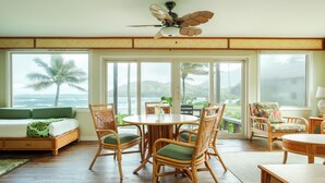 A spacious room with a large bed, a dining table, and chairs. There is a ceiling fan and large windows with a view of palm trees and mountains.