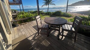 A wooden deck with a round table and chairs, overlooking a beach with palm trees and ocean waves.