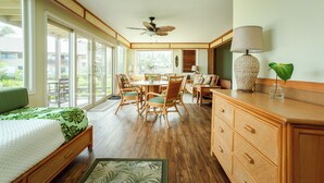 A spacious room with a bed, a dining table, chairs, a ceiling fan, and a large window providing a view of the outdoors.
