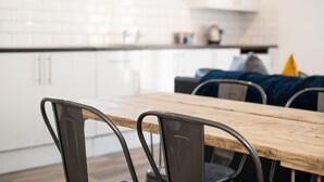 A dining area with a wooden table and metal chairs, white cabinets in the background, and a blue cushion on a sofa. - The Nook Close to Swansea Marina (Swansea)
