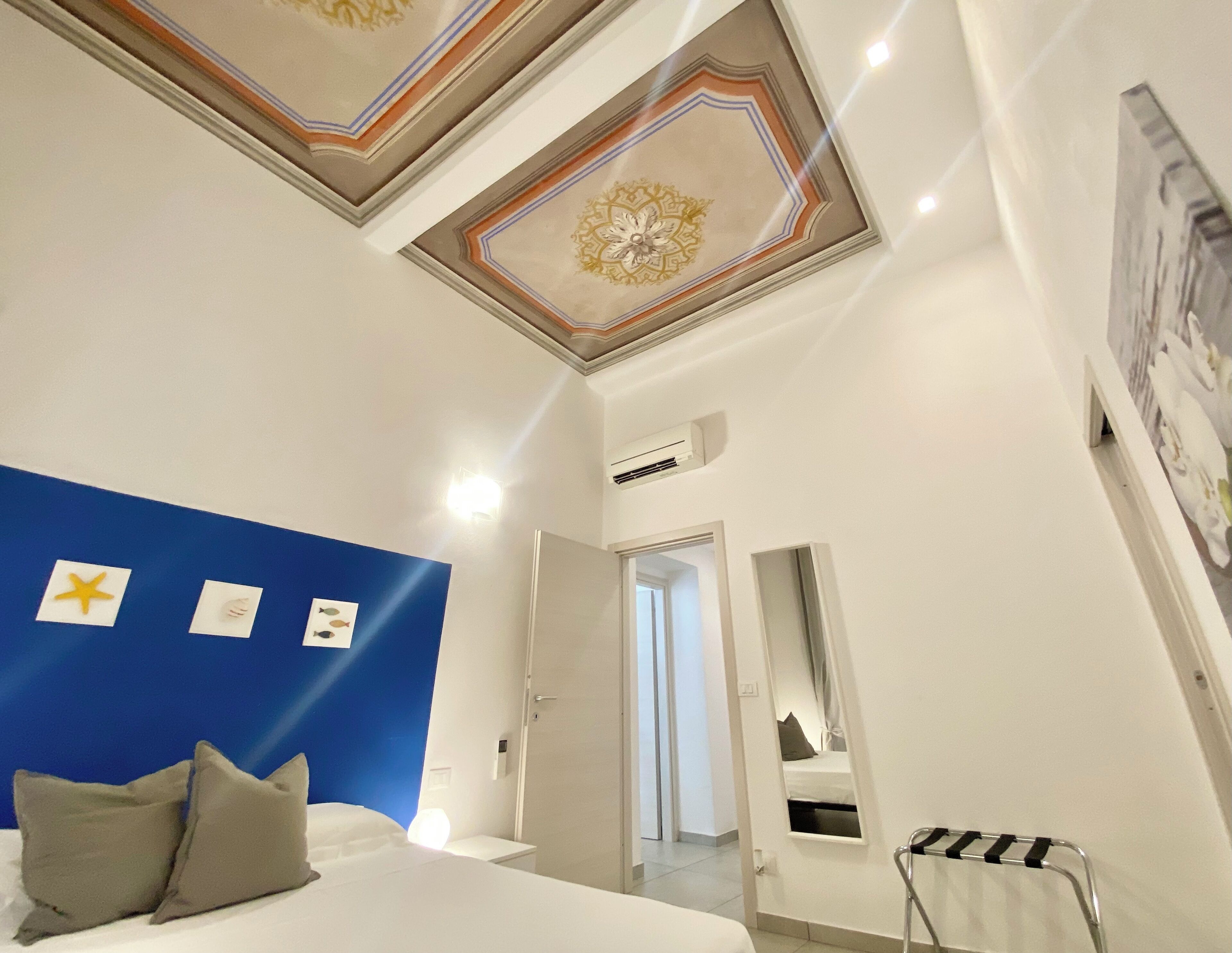 A hotel room with a blue accent wall, a bed with pillows, a ceiling with decorative patterns, and a mirror reflecting the room.