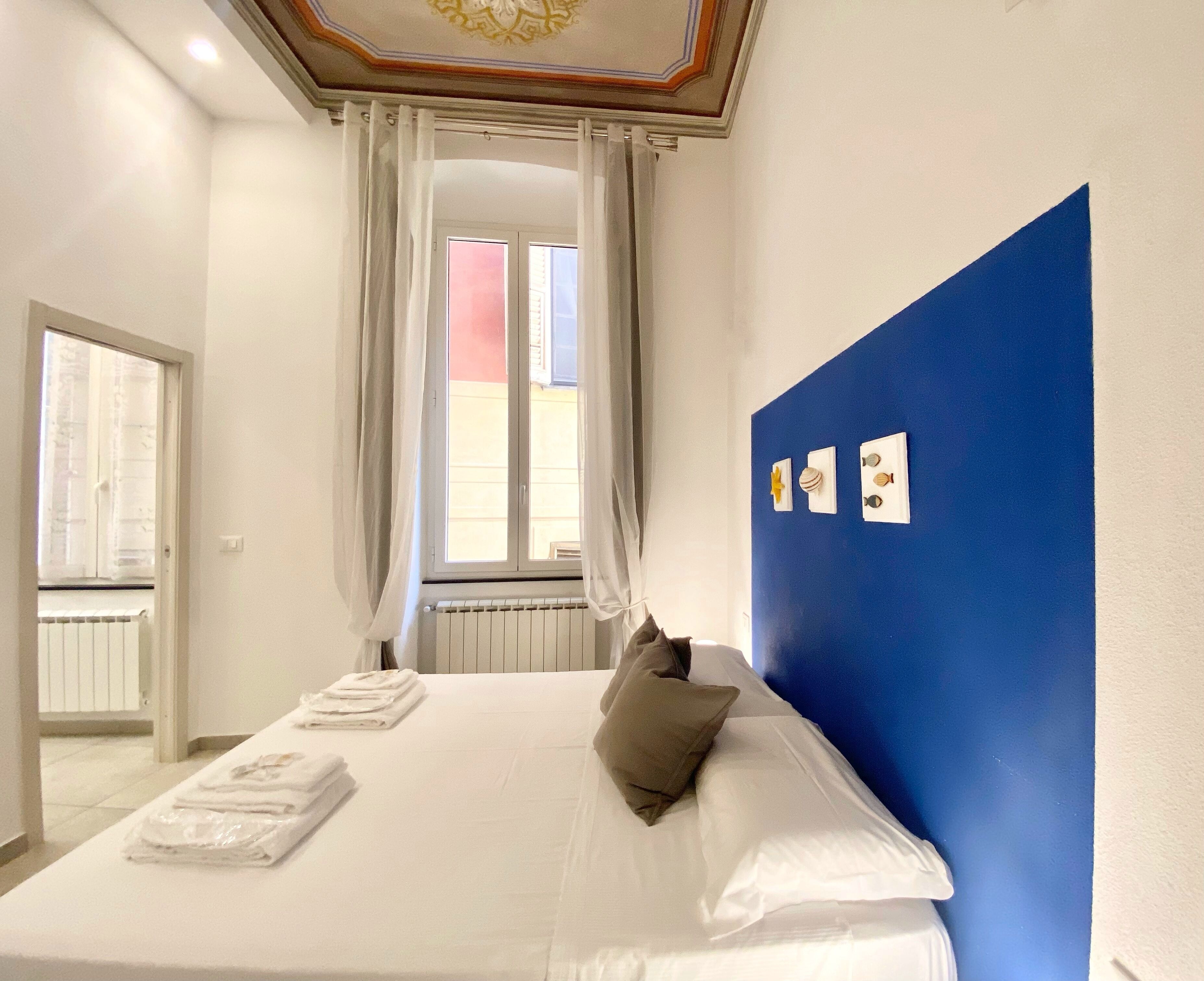 A hotel room with a bed, a window with curtains, a radiator, and a blue accent wall with wall art.