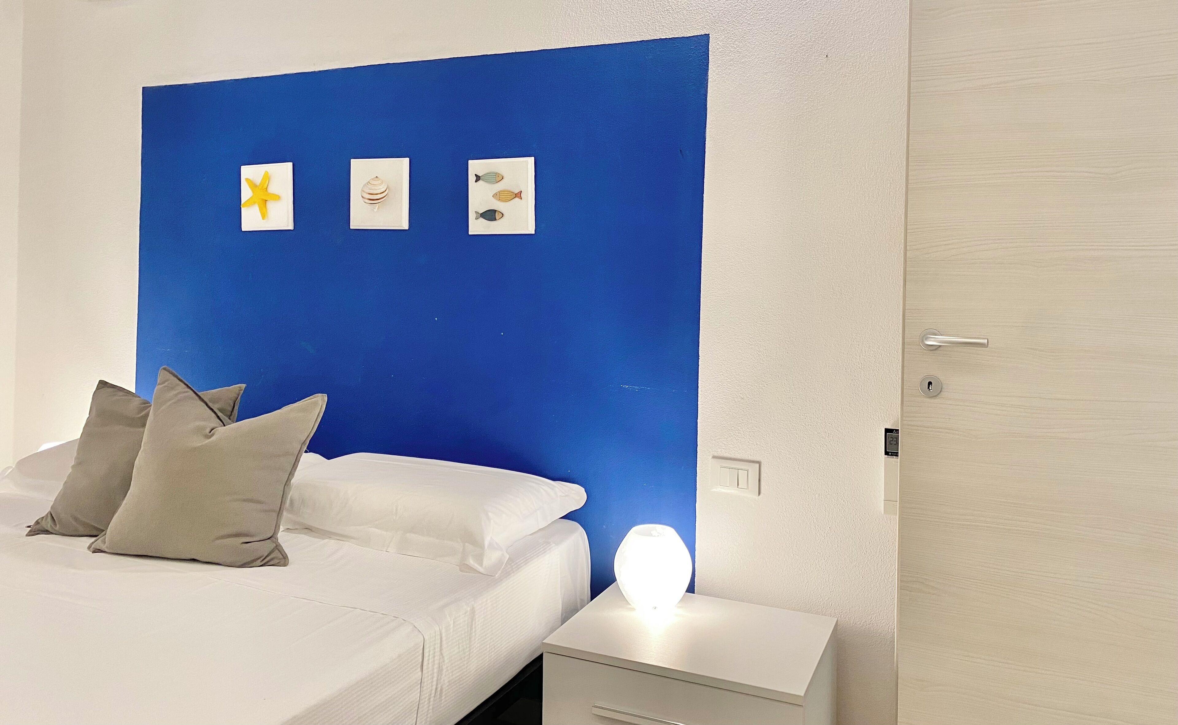 A bedroom with a blue accent wall, a bed with white bedding, two pillows, a bedside table with a lamp, and a wall-mounted light switch.