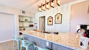 A kitchen with a granite countertop, two bar stools, a coffee maker, and two framed signs on the wall.