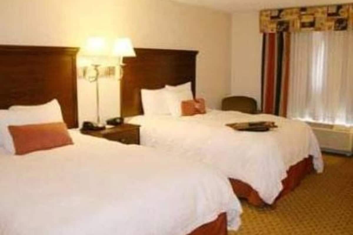 A hotel room with two beds, a nightstand, a lamp, and a window with curtains.
