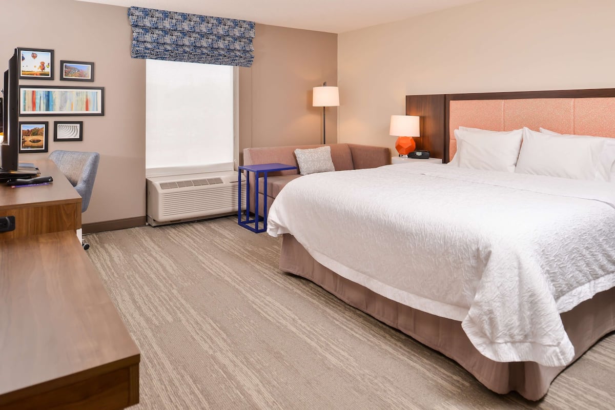 A hotel room with a large bed, a desk, a chair, a lamp, and a wall-mounted air conditioner.