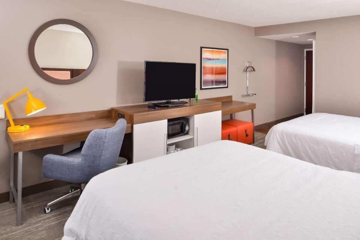 A hotel room with a desk, a chair, a television, a bed, and a small orange ottoman.