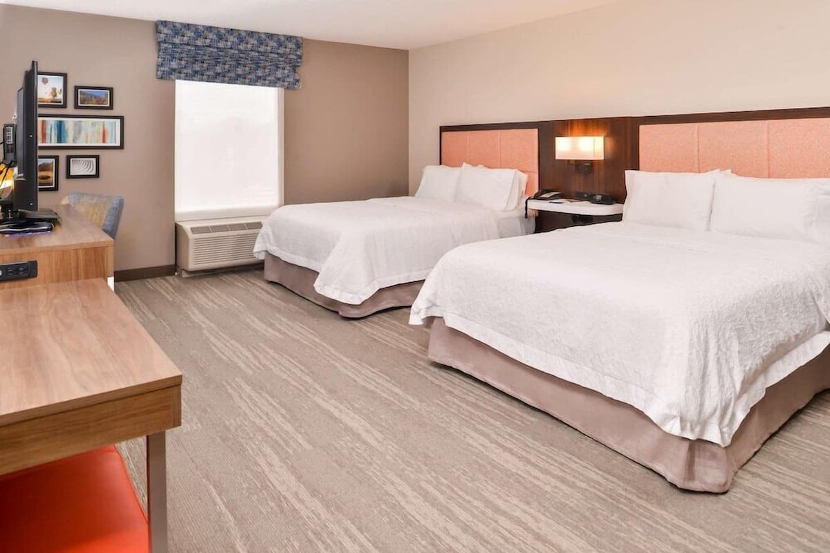 A hotel room with two beds, a desk, a chair, a TV, and a window.