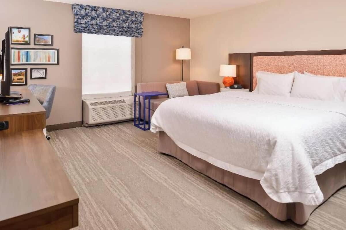 A hotel room with a large bed, a desk with a chair, a sofa, a standing lamp, and a wall-mounted TV.