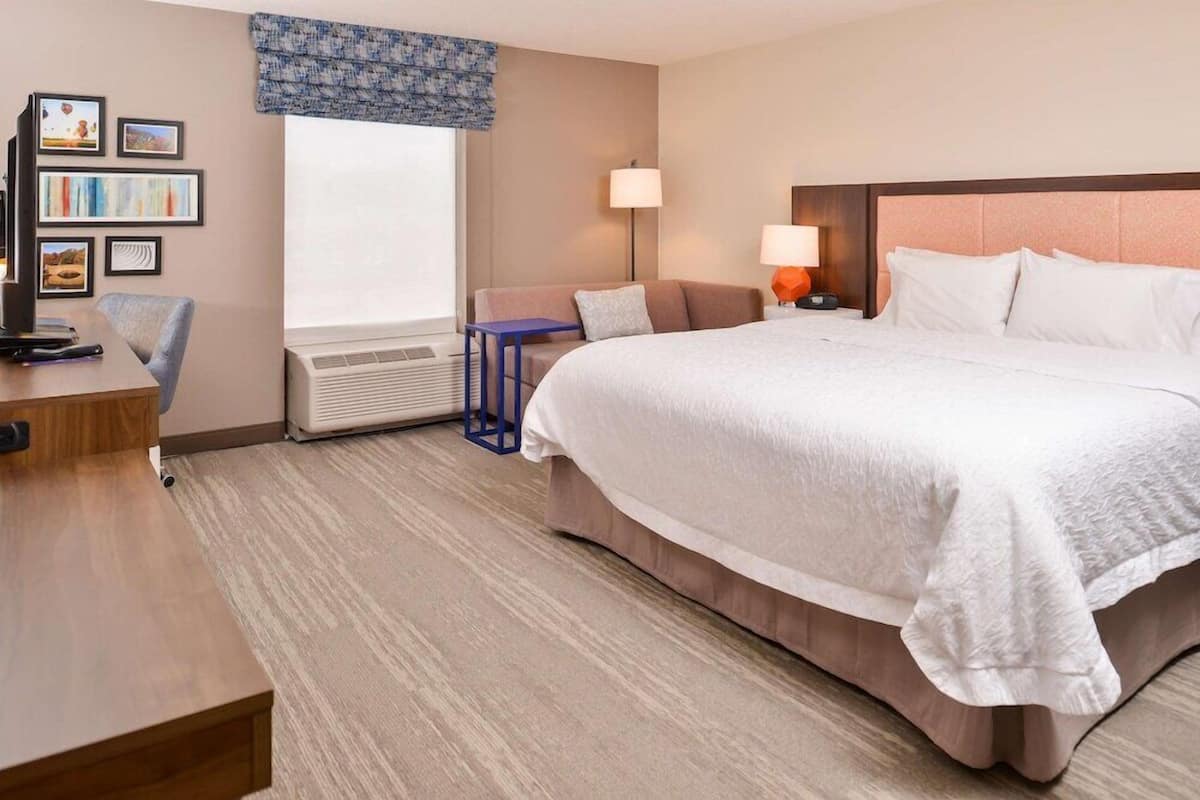 A hotel room with a bed, a desk, a chair, a lamp, and a wall-mounted TV.