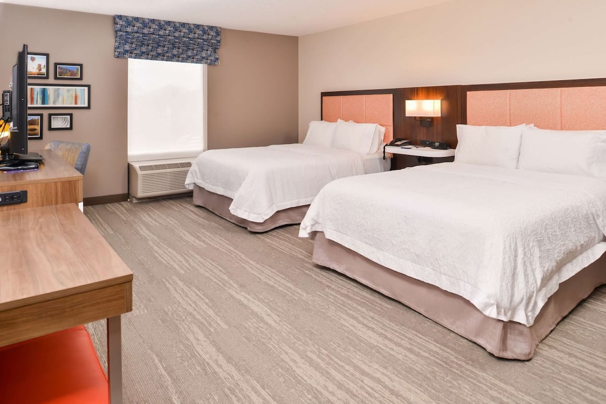 A hotel room with two beds, a desk, a chair, a TV, and a window.