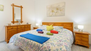 Een slaapkamer met een bloemenbedekking, een houten hoofdbord, een nachtkastje met een lamp en een houten dressoir met een spiegel.