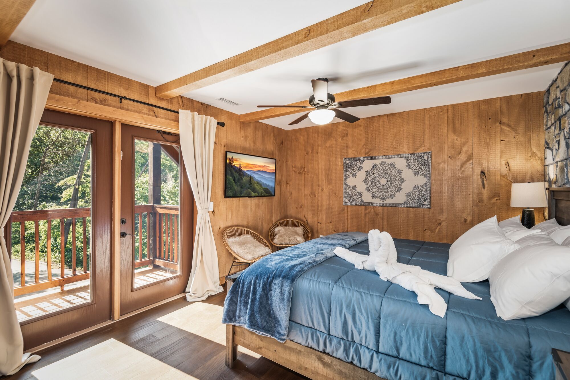 A bedroom with a wooden ceiling, a bed with blue bedding, a ceiling fan, a window with curtains, and a view of the outdoors.