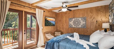 A bedroom with a wooden ceiling, a bed with blue bedding, a ceiling fan, a window with curtains, and a view of the outdoors.
