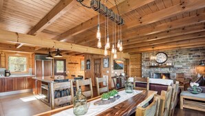 Interior rústico de una cabaña con una mesa de comedor de madera, sillas, una zona de cocina y una chimenea de piedra.
