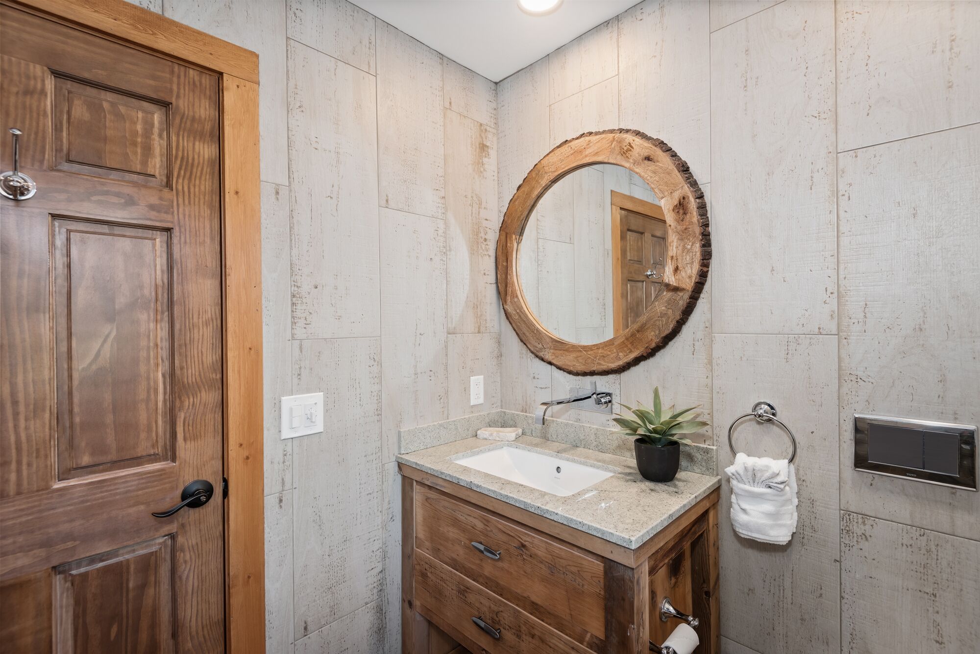 Un baño con puerta de madera, un espejo redondo, un lavabo con encimera de granito, un perchero y un dispensador de papel higiénico.