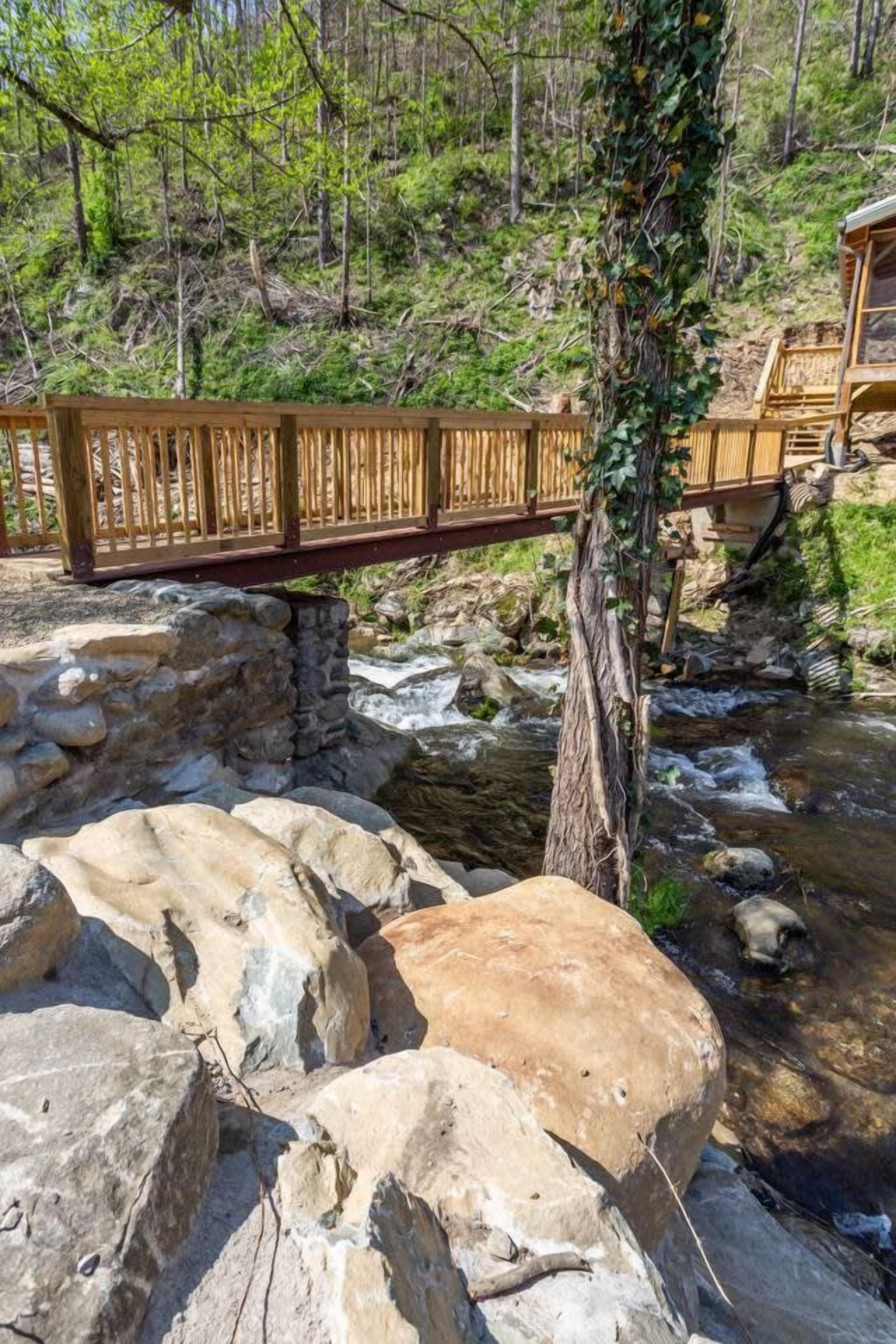 Un pont en bois enjambant un ruisseau, avec une structure en pierre en dessous, le tout entouré d’arbres et de verdure.