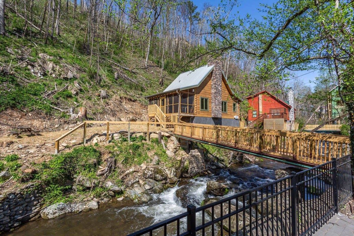 Eine Holzhütte mit einer Holzdar Terrasse und einem Steinkamin, gelegen an einem plätschernden Bach mit einer Holzbrücke, die darauf führt.