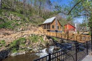 A cabin with a wooden deck and a stone chimney, situated by a flowing stream with a wooden bridge leading to it.