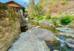 A stone wall with a wooden cabin, a small stream, and surrounding greenery.