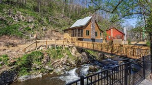 A cabin with a wooden deck and a stone chimney, situated beside a flowing river and surrounded by trees.