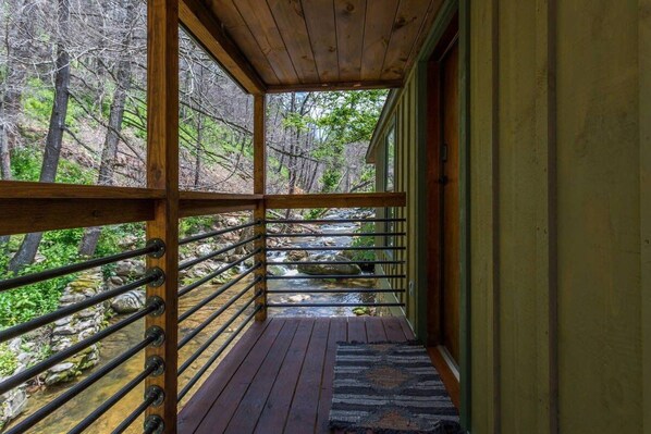 A wooden deck with a metal railing overlooking a stream and forest.