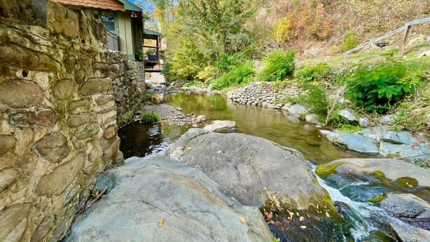 A rustic stone building adjacent to a flowing stream with a wooden deck and a view of a forested hillside.