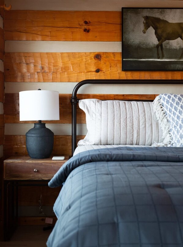 Un dormitorio con cabecera de madera, cama con colcha azul, mesita de noche con lámpara y un cuadro enmarcado de un caballo en la pared.