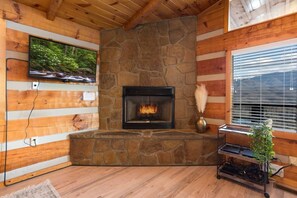 Una chimenea de piedra con fuego, un televisor montado en la pared y una ventana con persianas.