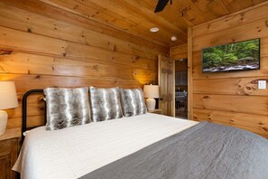 A wooden cabin bedroom with a bed, two wall-mounted lamps, and a television on the wall.