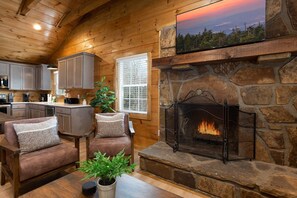 A cozy cabin interior with a stone fireplace, wooden walls, a kitchen area, and seating.
