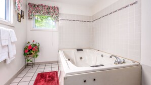 A bathroom with a white bathtub, a red rug, a window with floral curtains, and a potted plant with red flowers. - Parkway Inn (Waynesville)
