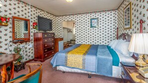 A bedroom with floral wallpaper, a bed with a blue and yellow quilt, a wooden dresser, a television, and a desk with a chair.