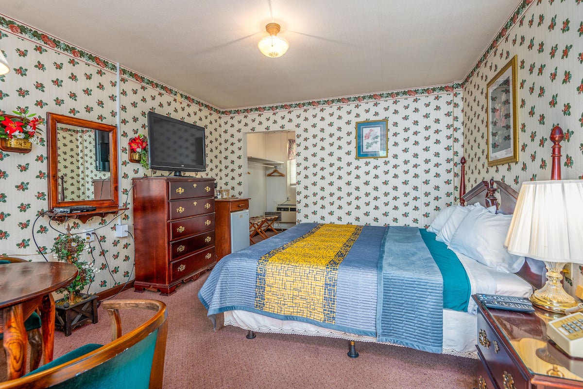 A bedroom with floral wallpaper, a bed with a blue and yellow quilt, a wooden dresser, a television, and a desk with a chair.
